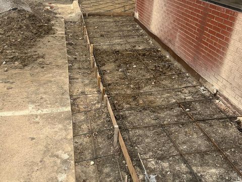 Uneven walkway under construction beside a brick wall, with exposed rebar and dirt trench.
