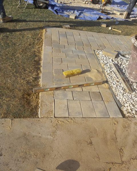 Broken walkway with loose pavers and a broom on a patio beside gravel and grass.