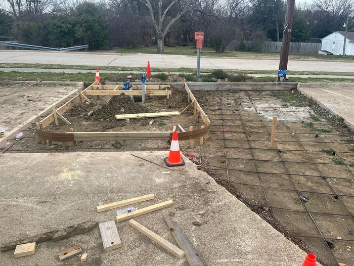 Construction site with trench excavation, orange cones, and wooden forms beside a driveway.