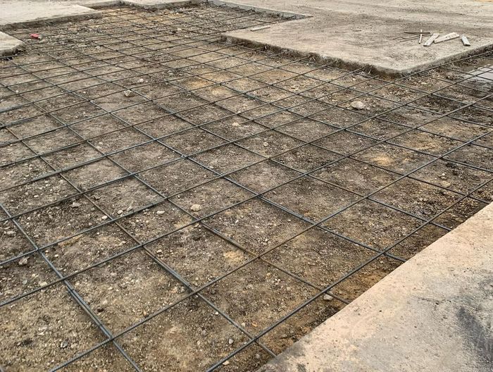 Rebar grid laid over a gravel base beside a concrete slab, preparing for construction