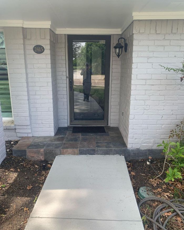 Front porch with glass door, brick walls, and a concrete walkway leading to the entrance.
