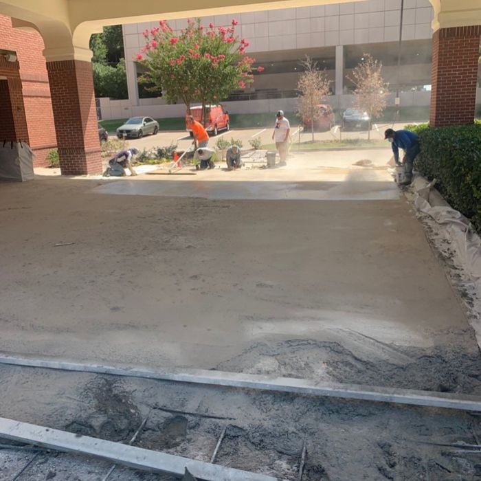 Workers smoothing fresh concrete under a covered walkway near a brick building and flowering tree