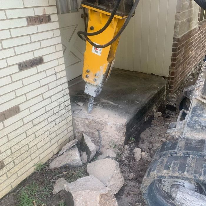 Excavator jackhammer breaking concrete beside a brick wall and doorway, with rubble on the ground.