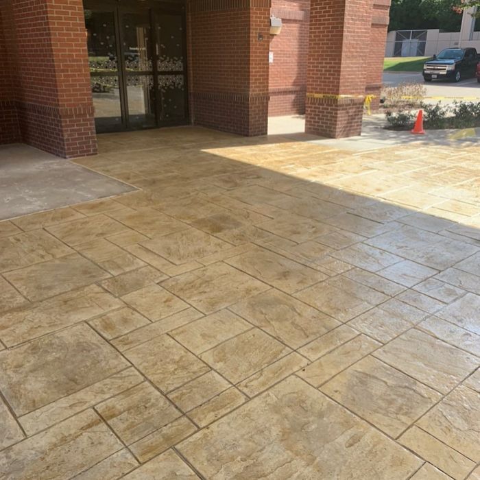 Stone-patterned walkway outside a brick building entrance, with sunlight and a parking area visible nearby