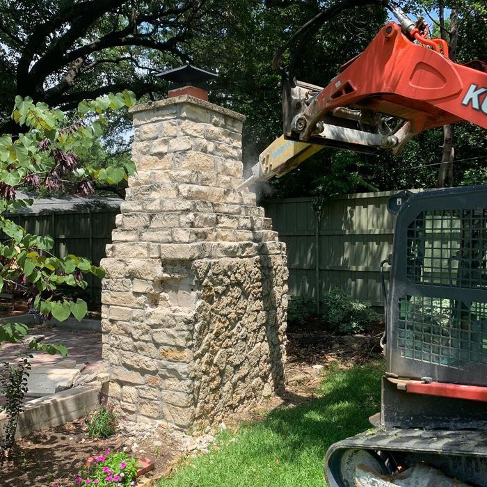 Excavator arm beside a stone chimney in a leafy backyard, with a green fence in the background
