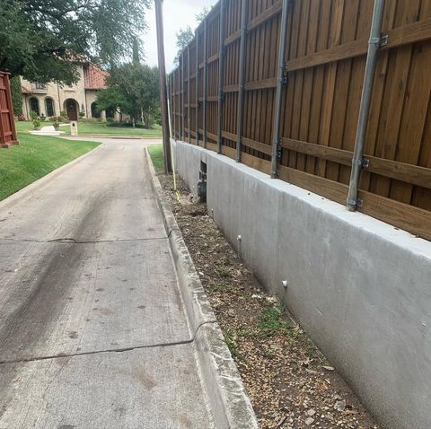 Narrow concrete sidewalk beside a wooden fence, leading past a grassy courtyard with buildings in the distance