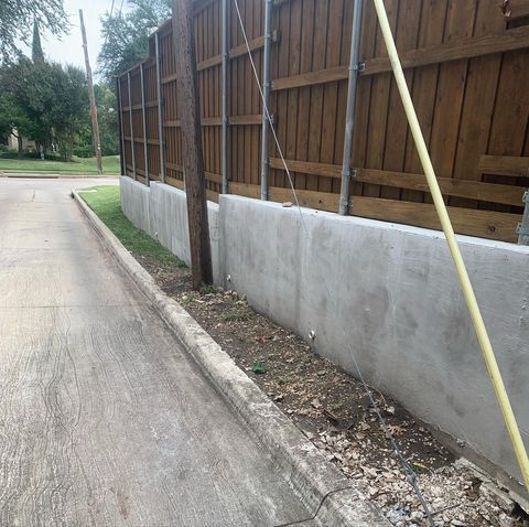 Concrete roadside wall beneath a wooden fence along a quiet street, with poles leaning against it.