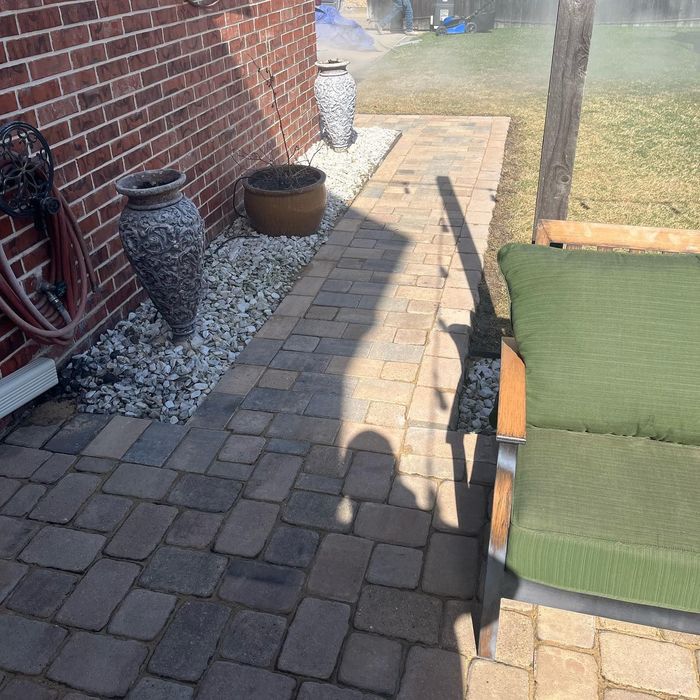 Brick patio beside a house with pavers, large planters, and a green chair in sunlight.