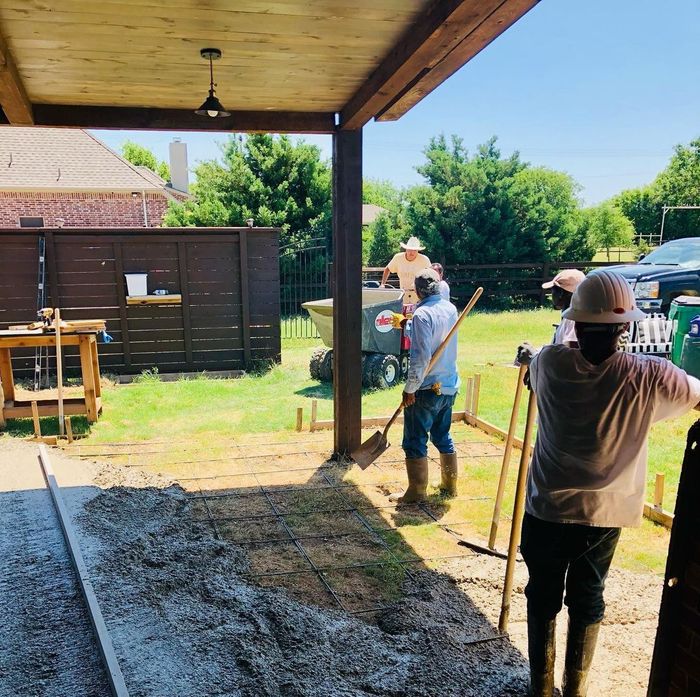 Workers leveling concrete in a shaded backyard patio area with tools and gravel.