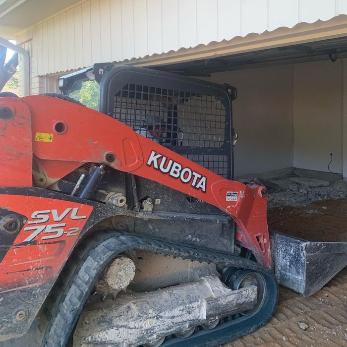 Red Kubota skid steer half inside a shed, pushing debris from a doorway onto a muddy floor.