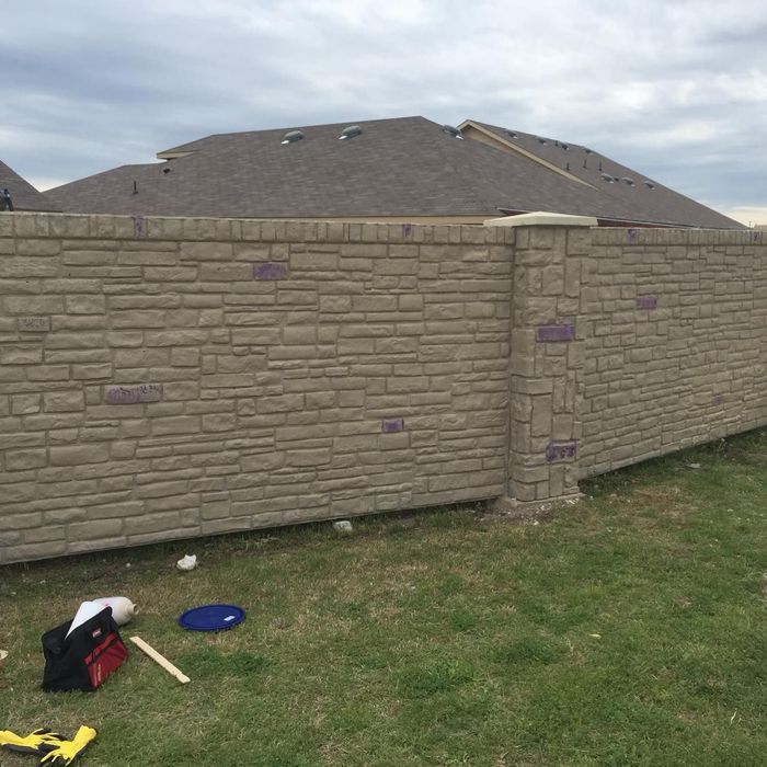 Backyard with tan brick wall, house roof in background, and lawn tools on the grass.