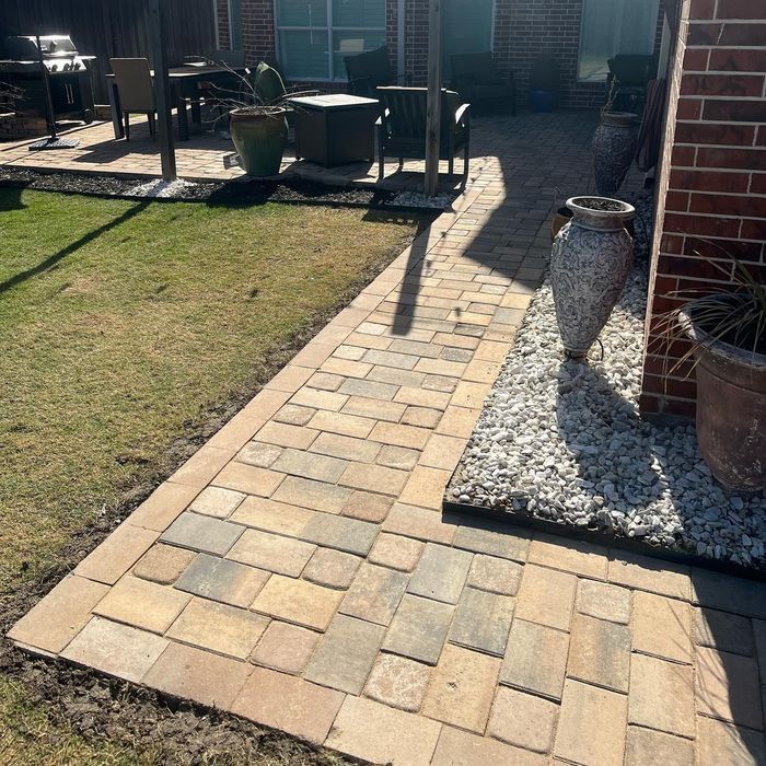 Paver walkway beside a brick house, bordered by gravel, with a grassy patio area and outdoor furniture.