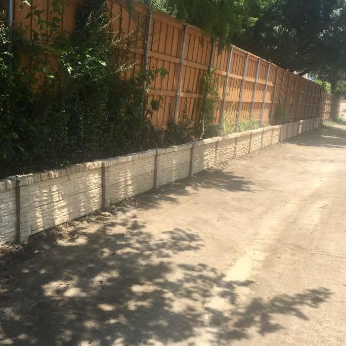 Sunlit dirt road beside a low white wall and tall wooden fence, with tree shadows across the path