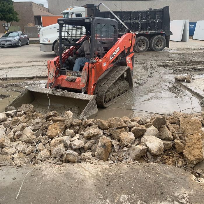 Orange skid steer pushing rocks through muddy construction site, with dump truck in background