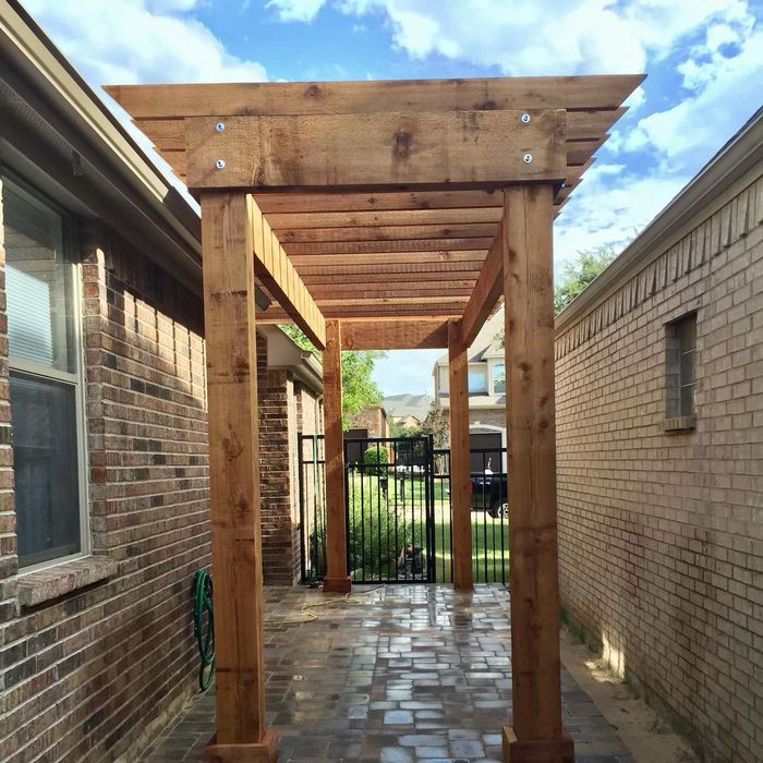 Wooden pergola walkway between brick buildings, leading to a black gate and grassy yard.