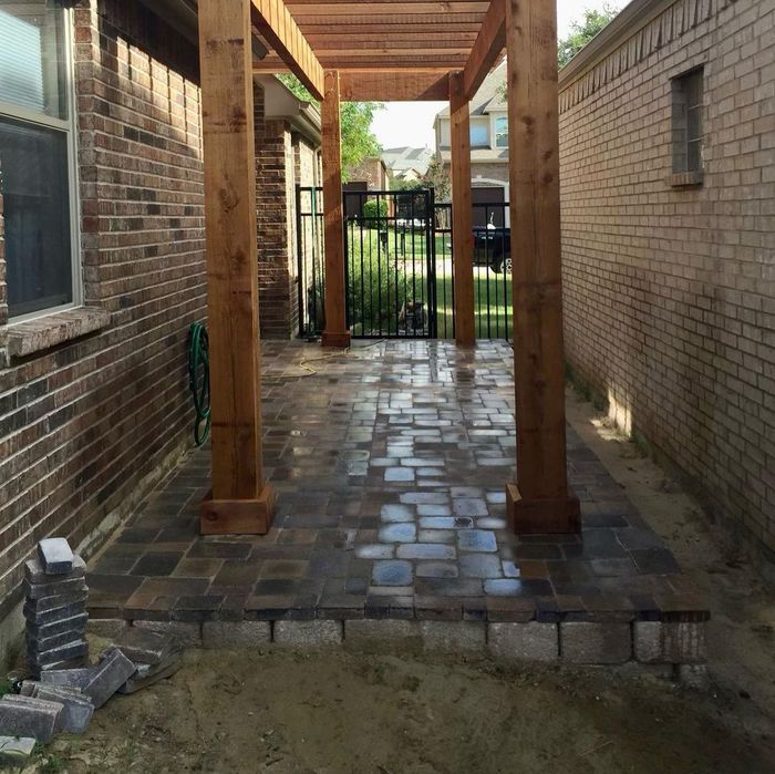 Brick courtyard walkway with wooden posts between brick walls, leading to a black gate and grassy yard.