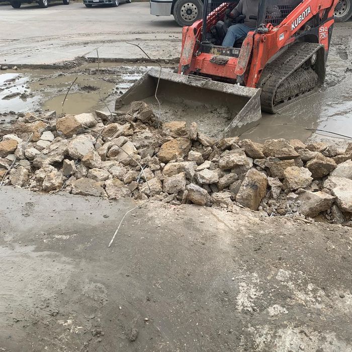 Orange skid steer pushing rocks and debris on a wet street near a broken curb