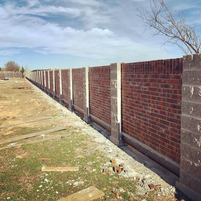 Long brick wall with concrete posts along a dry, open field under a cloudy sky
