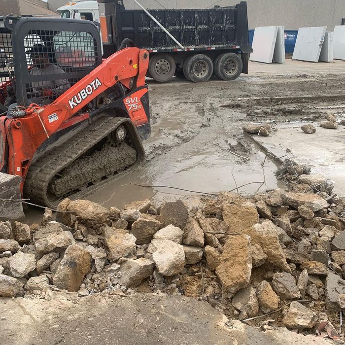 Orange Kubota skid steer pushing rocks and mud on a construction site near a dump truck.