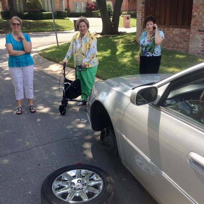 Three people stand by a silver car with a flat front tire and detached wheel on a driveway.
