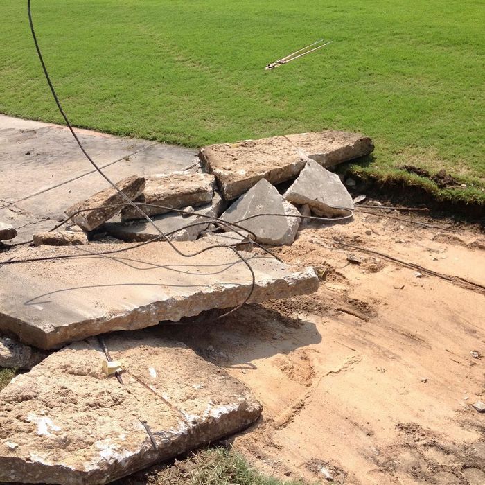 Broken concrete slabs and exposed wires at the edge of a grassy lawn, showing ground collapse.