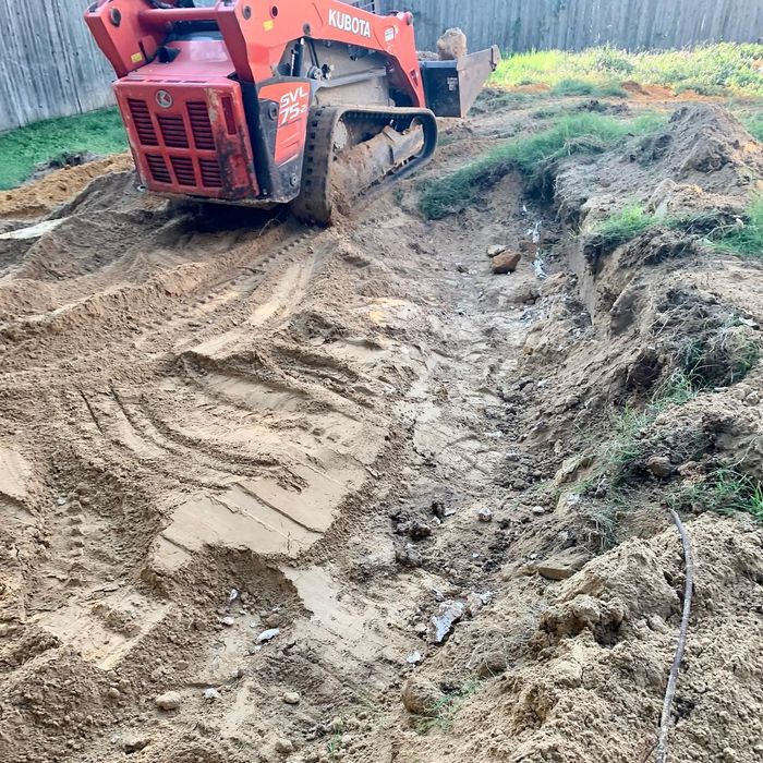 Red excavator grading a muddy dirt path beside a grassy embankment