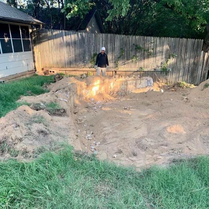 Worker in a backyard excavation site beside a house and wooden fence, with exposed dirt and a lit fire pit.
