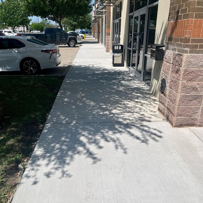 Sunny sidewalk beside storefronts with parked cars and tree shadows.