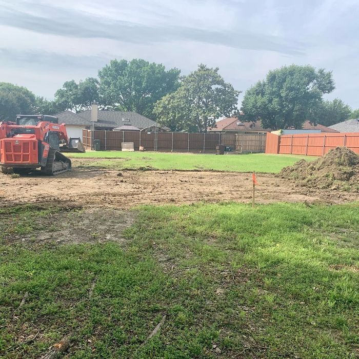 Backyard under construction with a skid steer, dirt pile, and wooden fence under a sunny sky