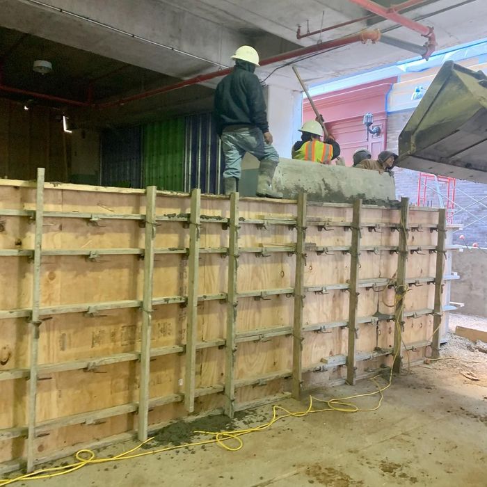 Construction workers pouring concrete into a large wooden form inside an indoor site.