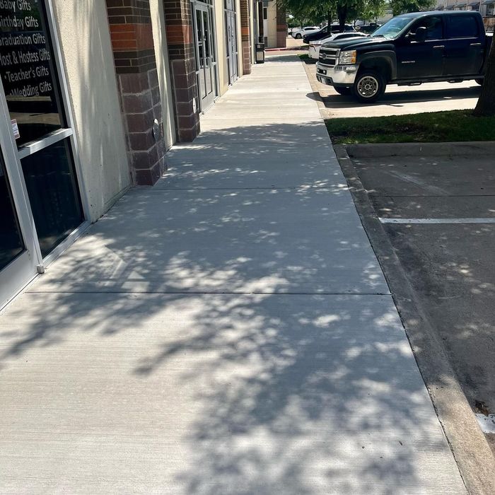 Sunlit concrete sidewalk beside a building, with parked black truck and tree shadows.