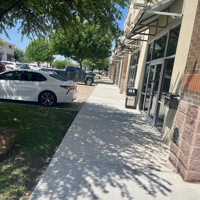 Sidewalk outside storefronts with parked cars and tree shade on a sunny day