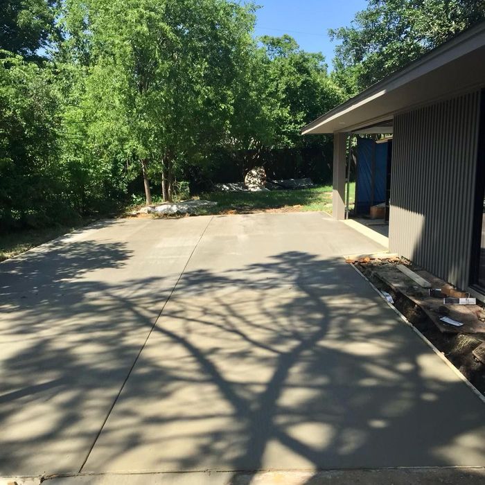 Concrete driveway beside a dark-sided house, with trees and sunlight casting shadows.