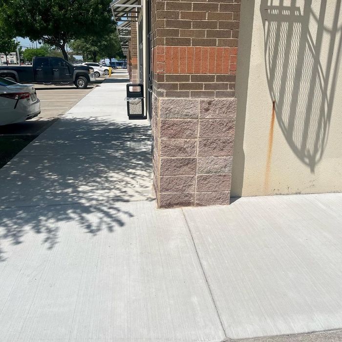 Sidewalk beside a brick-and-stone building with parked cars and tree shade in the background