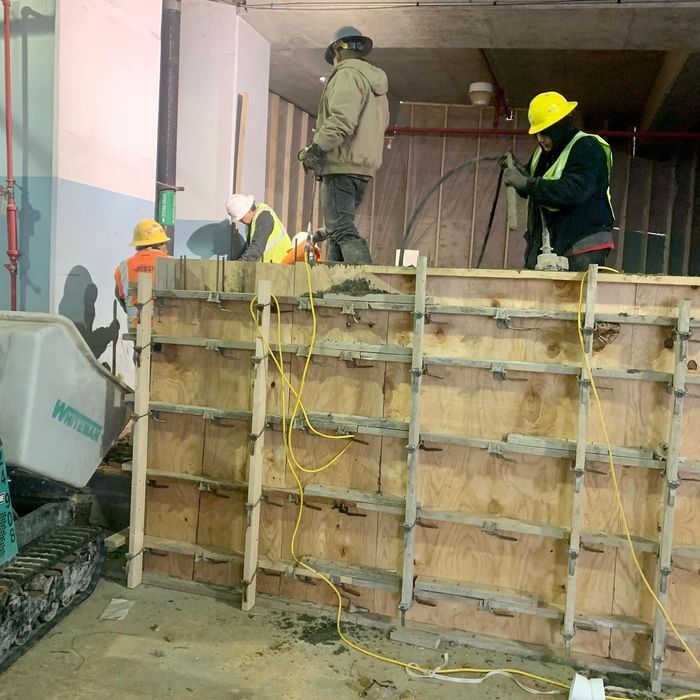 Construction workers on a wooden formwork wall in a dim indoor site, one wearing a yellow hard hat.