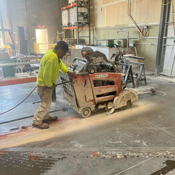 Worker using a floor saw in a dusty workshop, cutting a line into the concrete floor.