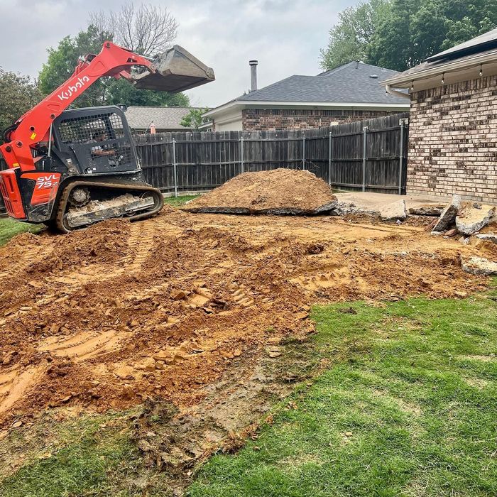 Skid steer leveling dirt in a backyard beside a fence and house during landscaping work