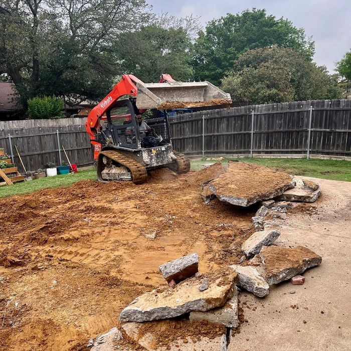 Orange excavator clearing dirt and broken concrete in a fenced backyard.