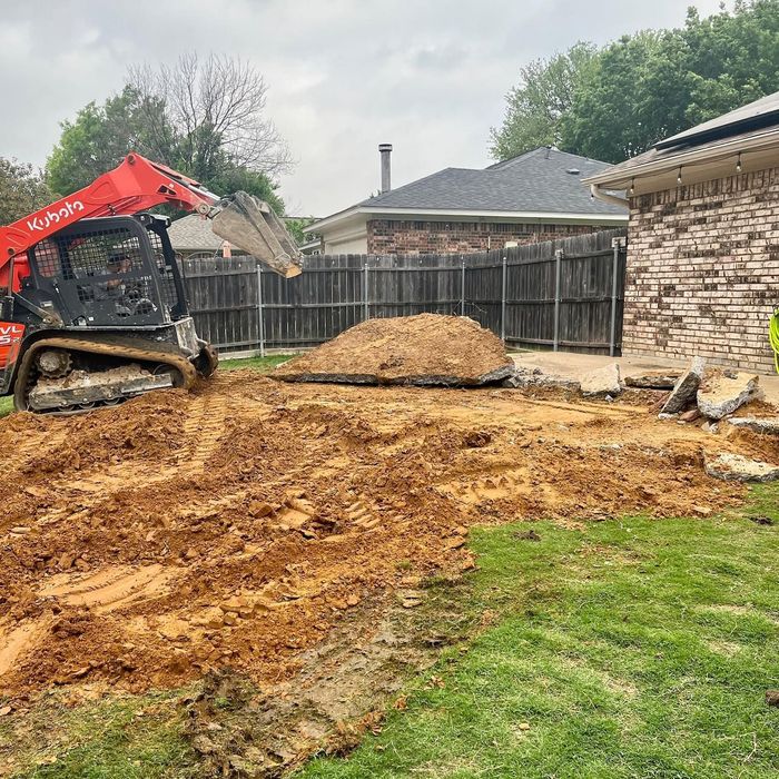 Mini excavator leveling dirt in a backyard beside a brick house and fenced yard