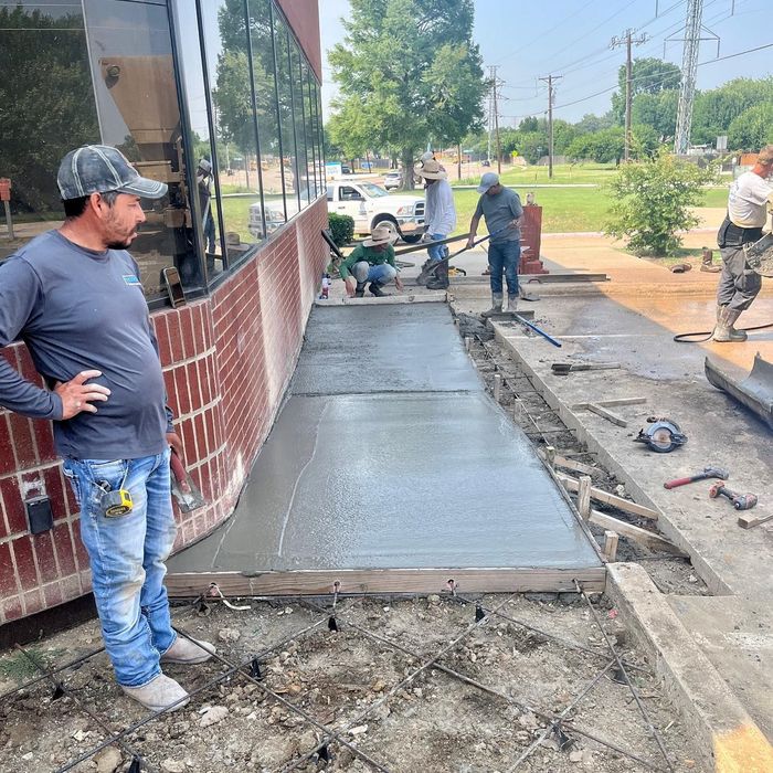 Workers pouring a new concrete sidewalk beside a brick building on a sunny day