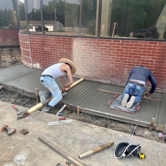 Two workers installing concrete pavers beside a curved brick building.