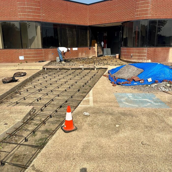 Construction site with rebar slab, blue tarp, and orange cone in front of a brick building.