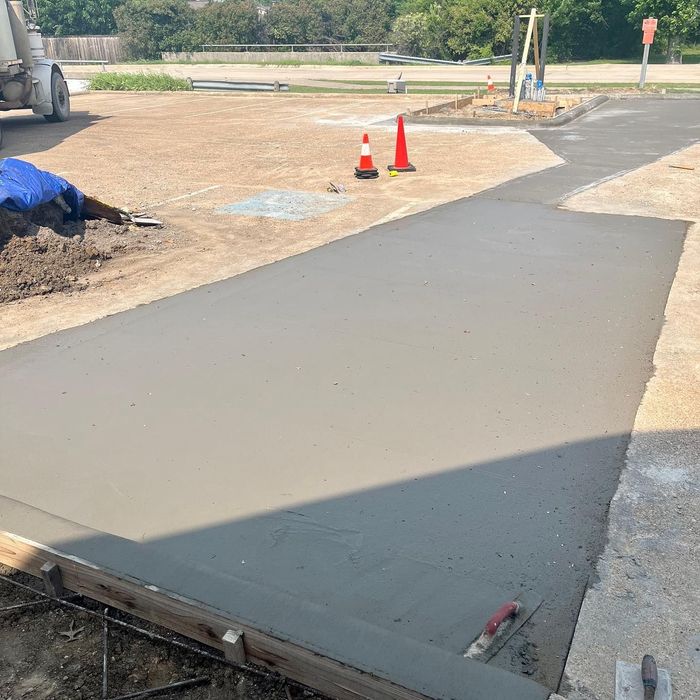 Freshly poured concrete walkway beside a road, marked by two orange traffic cones.
