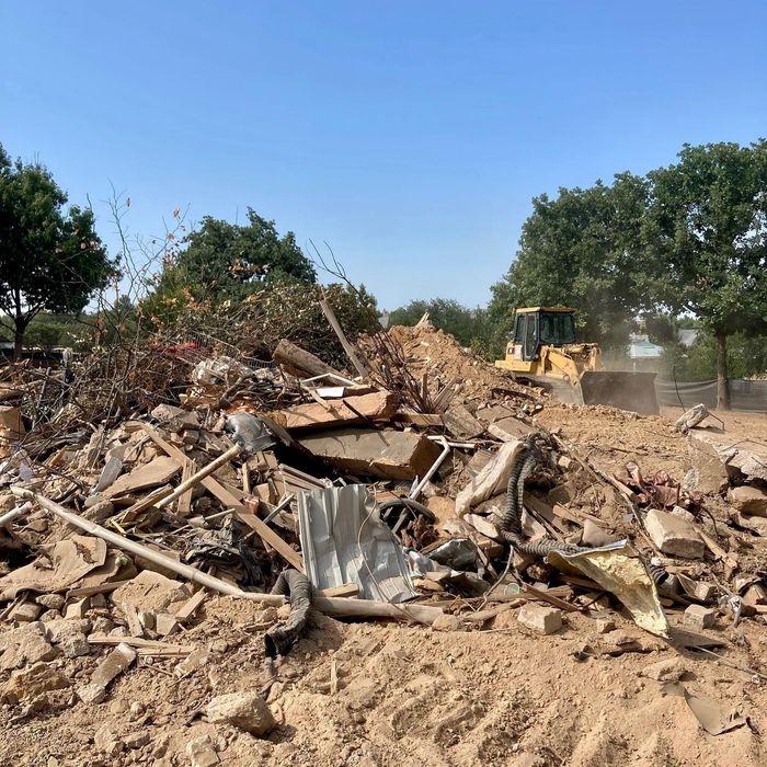 Pile of demolition debris and dirt in a cleared lot, with trees and a bulldozer in the background.