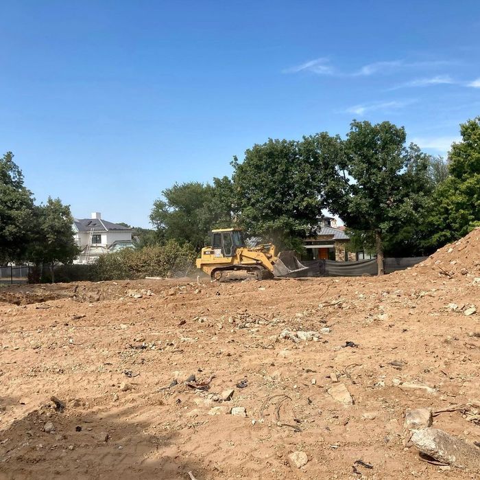 Bulldozer clearing a dirt lot under a clear blue sky, with trees and houses in the background