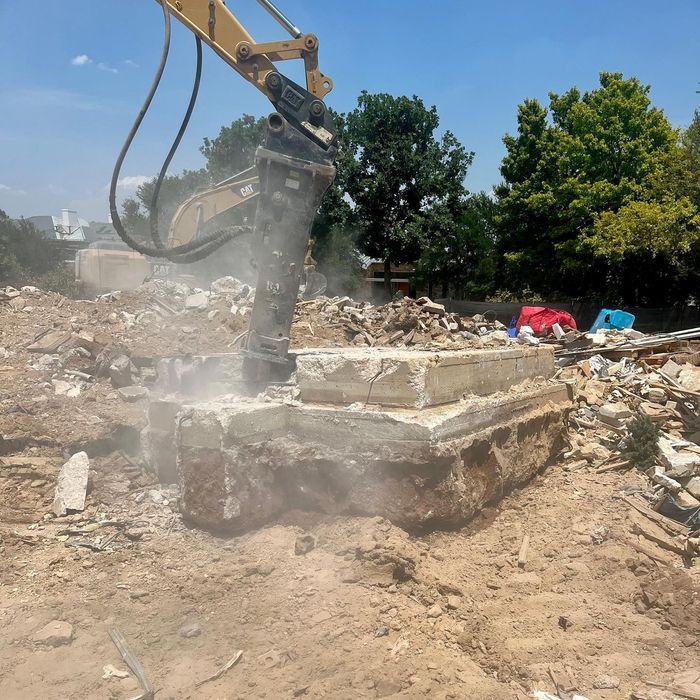 Excavator breaking concrete rubble at a dusty demolition site under clear blue sky.