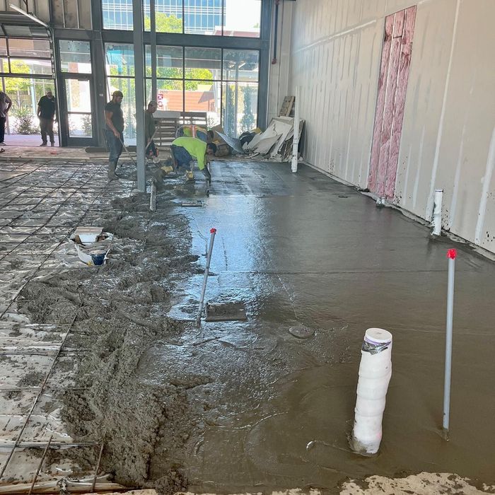 Construction site with wet concrete floor, workers in background, and a wall opening near glass doors.