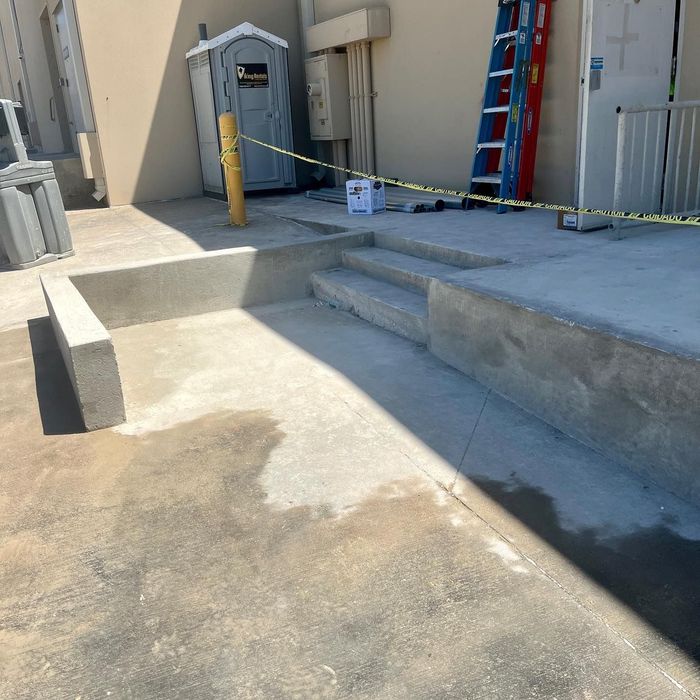 Outdoor utility area with concrete ramps, caution tape, ladder, and a portable toilet beside a building