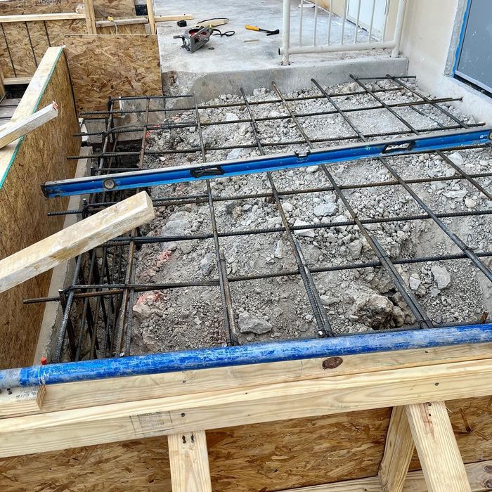 Construction site with rebar grid over concrete, framed by wood and blue pipes near a doorway