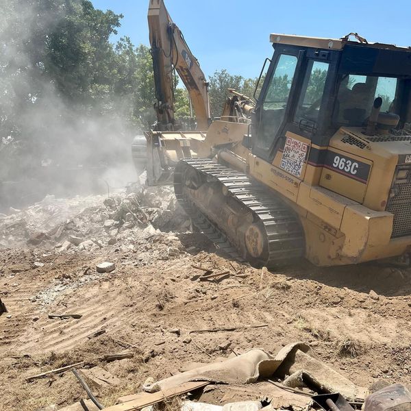 Yellow excavator demolishing rubble, kicking up dust at a construction site